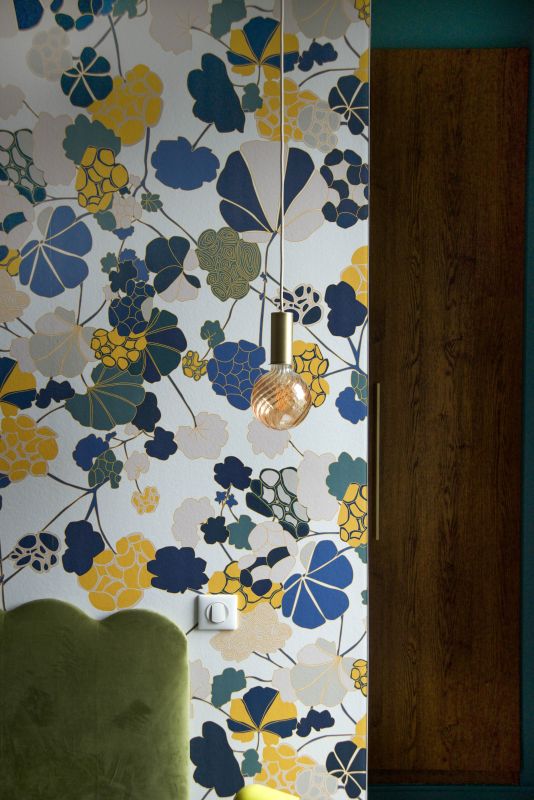 détail sur un papier peint de chez Ressource dans les tons de bleu, jaune, vert et blanc, suspension laiton Collect de chez Ferm Living, tête de lit en velour et en arrière plan des façades de dressing en noyer et une poignée en laiton de chez LMC