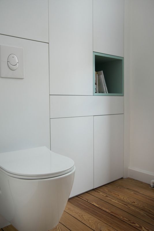 il s'agit d'un wc, avec une cuvette suspendue, le bâti support est intégré dans un meuble sur mesure blanc comprenant une niche vert d'eau avec des livres, il y a du parquet ancien