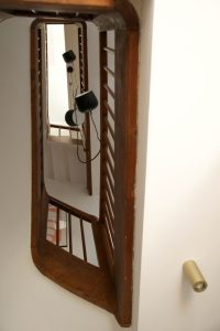 photo d'une cage d'escalier vue en contre plongée, garde-corps en bois et suspension noire Aim de chez Flos, les plafonds sont blancs et on aperçoit en spot en saillie en laiton de chez SLV