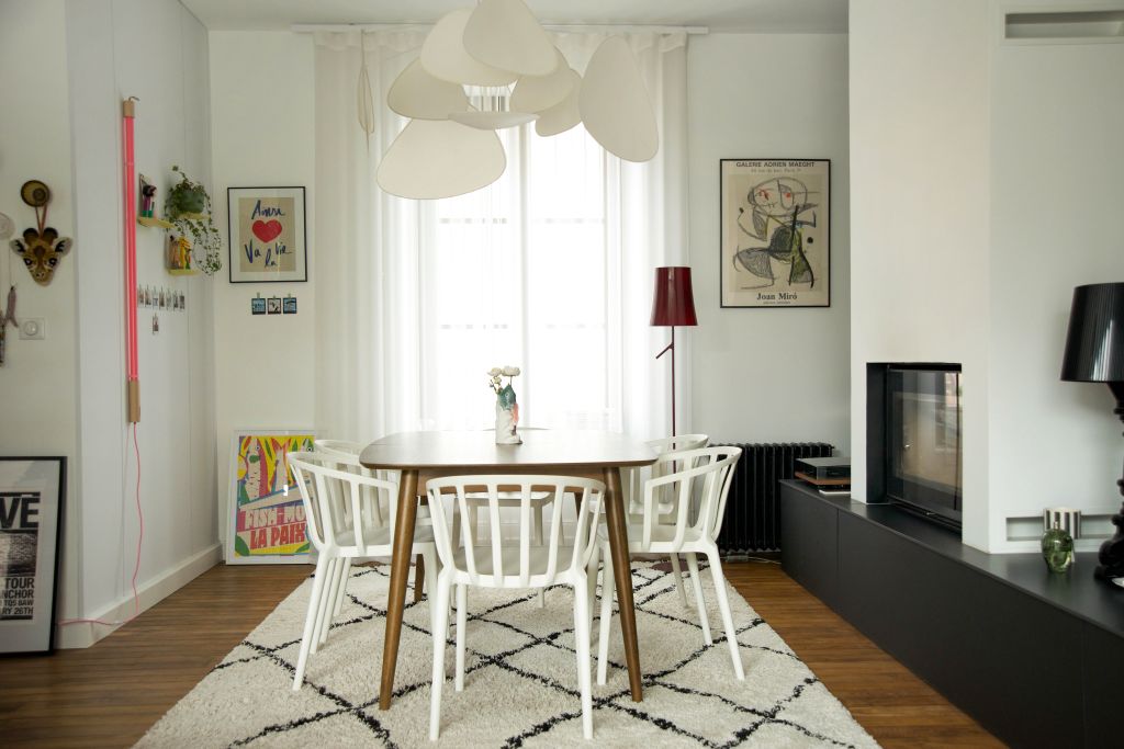 photo d'une salle à manger comprenant une table en noyer et des chaises blanches Venice de chez Kartell, tapis berbère noir et blanc, d'une suspension blanche Screen de chez Market Set, il y a du contraste, une t une lampe Birdie de chez Foscarini de chez artemide, revêtement mural noir Facet de chez Arte, cheminée encastrée Heta sur banc noir avec tiroirs, néon rose fluo de chez Seletti, des étageres jaunes des chez Moustache, des affiches colorées