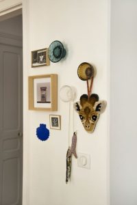 zoom sur un détail avec des décorations colorées suspendues (3 cadres avec photos, une coquille saint jacques bleu yves Klein de chez Monochromic Ceramic, sardines en tissu...) sur un mur blanc ainsi que des patères Jellies Familyde chez Kartell