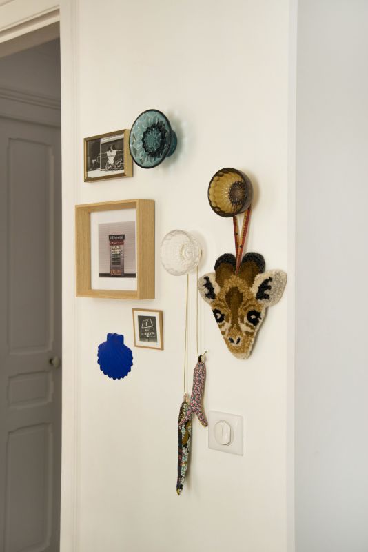 zoom sur un détail avec des décorations colorées suspendues (3 cadres avec photos, une coquille saint jacques bleu yves Klein de chez Monochromic Ceramic, sardines en tissu...) sur un mur blanc ainsi que des patères Jellies Familyde chez Kartell