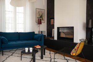 photo d'un salon avec un canapé bleu en laine bouillie, un tapis berbère, des tables noires T-table de chez kartell avec des mugs de chez Felicie aussi, un fauteuil noir en métal Hay, d'une suspension blanche Screen de chez Market Set, il y a du contraste, une niche avec des vinyles et une lampe Birdie de chez Foscarini de chez artemide, revêtement mural noir Facet de chez Arte, cheminée encastrée Heta sur banc noir avec tiroirs