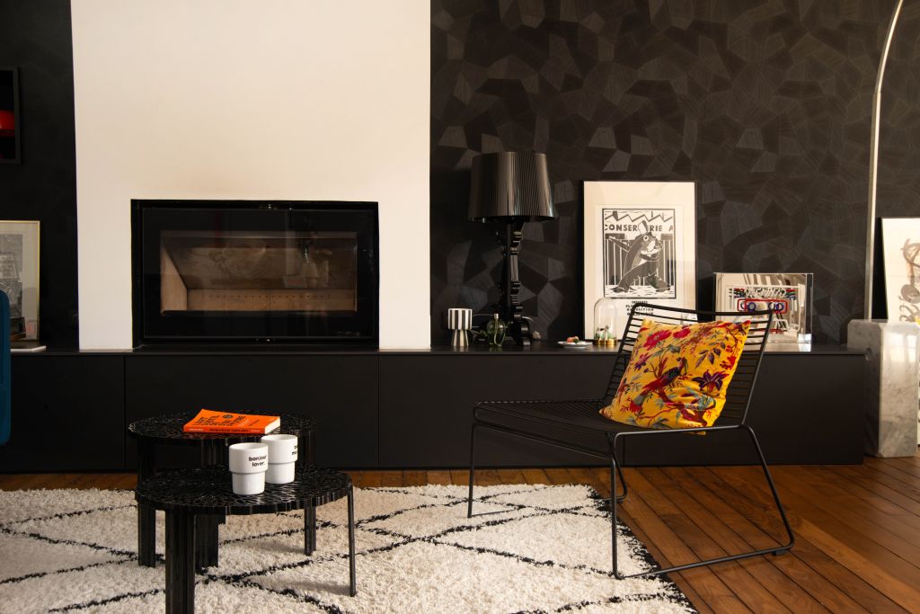 il s'agit d'un espace salon majoritairement noir et blanc, on peut y voir des fauteuils noirs en métal Hay, des tables noires Kartell, un tapis berbère blanc, une cheminée encastrée Heta posée sur un banc réalisé sur mesure en céramique et mélaniné. Il y a du parquet ancien et un revêtement mural noir de chez ARTE.